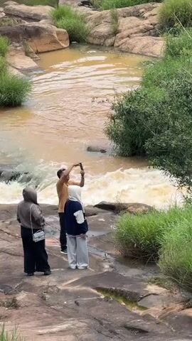 The video shows a rocky, natural setting with a flowing river. In the foreground, three people are standing on rocks, two women and one man. The man is holding up a phone, possibly taking a picture or video. One woman is wearing a grey hijab and dark clothing, and the other is wearing a white hijab and a blue and white top. Further in the background, two more women are visible. One is wearing a red dress and is sitting on a rock, while the other, wearing a patterned jacket and skirt, is standing. The river is a muddy brown color, indicating it may be carrying sediment. Lush green vegetation is abundant throughout the scene, with trees and tall grass growing along the riverbanks and on the rocky terrain. The overall environment appears to be a natural landscape, possibly a park or a wilderness area.