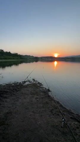 The video opens with a wide shot of a calm lake at sunset. The sun, a bright orange orb, is positioned near the horizon, casting a shimmering reflection on the water's surface. On the muddy bank in the foreground, two fishing rods are visible, their lines extending into the lake. The camera slowly pans to the right, revealing more of the tranquil lake and the distant tree-lined hills. The sky transitions from a soft orange near the horizon to a clear blue overhead. The serene atmosphere is accentuated by the gentle rippling of the water.