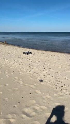 The video shows a person's hand reaching towards a small drone on a sandy beach. The drone takes off and hovers briefly before flying higher into the clear blue sky. In the background, the ocean meets the horizon. Three people, including what appear to be children, stand near the shoreline, watching the drone. The beach is sparsely populated, with some seaweed or debris scattered on the sand. The weather is sunny and calm.