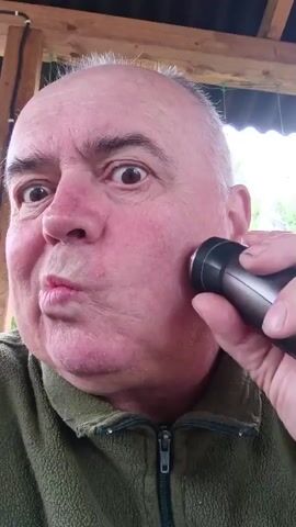 A close-up shot shows an older man with fair skin and graying hair as he shaves his face with an electric razor. He is wearing a green fleece jacket and is outdoors, with a wooden roof structure visible above him. The man is puckering his lips as he moves the razor across his cheek. The video is shot from a selfie perspective.