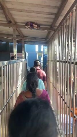 The video shows a crowded queue of people moving along a narrow, covered walkway bordered by metal railings on both sides. The walkway appears to be outdoors, with sunlight streaming in from the end of the passage. The ceiling is made of corrugated material supported by wooden beams. Several individuals are visible in the queue, all facing away from the camera and moving forward. Their clothing suggests a warm climate. The overall atmosphere seems to be one of orderly movement within a public space, possibly a temple or a similar place of worship or gathering. The camera is positioned behind a person in the queue, looking forward down the line.