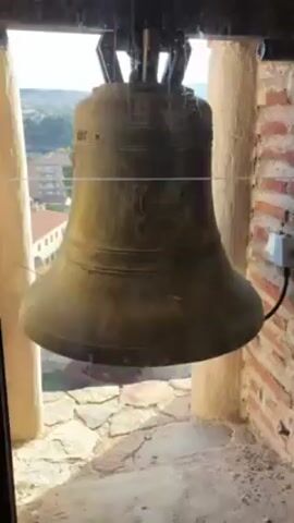The video shows a large church bell in a brick tower room. The bell is mounted high with dark metal framing and wiring visible. It is a dull, worn color, suggesting age. Outside the window, there is a view of rooftops and a distant hillside. The camera pans to show a narrow window with a dark frame and a stone and brick wall. A wooden railing is partially visible, leading to an unseen area below. The overall setting is a rustic tower with historical features.