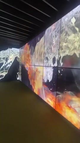 A wide-angle shot of a room with walls covered by a large video display. The display shows swirling abstract imagery of water and fire. The camera is positioned on a yellow-green floor and pans to the right. The video walls are made up of many individual screens.