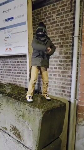 A young person stands atop a large, moss-covered concrete block in front of a brick wall with a white sign attached to it. The person wears a dark green coat, tan pants, a black beanie, and a dark neck gaiter pulled up to cover most of their face. Their sneakers are predominantly white with black and yellow accents. They initially hold one finger up in the air, then point to the right with that arm, while the other hand is on their hip. The sign on the brick wall lists figures in Euros for various projects: "Pas de Calais", "Hauts-de-France", and company details with listed prices, such as "BOUYGUES ENERGIES & SERVICES" and "DUJARDIN CONSTRUCTION". A white drainpipe runs vertically along the wall to the right of the concrete block. The setting appears to be an outdoor urban area at night, with the lighting primarily illuminating the subject and immediate surroundings.