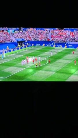 A soccer match is shown in a stadium with a large crowd. The field is green with white lines. Players in red and white uniforms are on the field. A referee in a yellow shirt is also present. The scoreboard shows the score as 0-0. The game is part of the FIFA World Cup 2026.