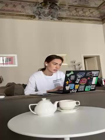 A woman with long brown hair is sitting on a gray couch, typing on a black laptop. She is wearing a white long-sleeved shirt and white earbuds. The laptop is covered in stickers. A white teapot and a white teacup and saucer are on a round white table in front of the couch. The room has light-colored walls and a decorative ceiling. A door is visible in the background. There is a framed picture on the wall behind the couch.