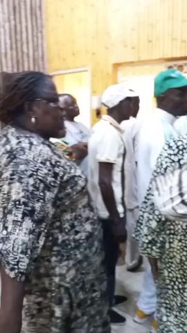 The video shows several people standing in an indoor space with a wooden wall, looking at two framed pictures hanging on the wall. One person with a green cap seems to be pointing at the picture. The people are wearing different clothing styles, including patterned shirts and white shirts. The scene appears to be in a house or an office space.