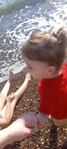 This is a short, shaky video clip taken from a first-person perspective at a pebble beach. The foreground is dominated by the hand of the videographer, holding the hand of a small child. The child, wearing a red shirt, is looking off to the left of the frame. Behind the child and the videographer's legs are the bare legs and feet of another person, lying on the pebbles. The sea is visible in the upper part of the frame, with waves breaking on the shore. The scene is lit by bright sunlight, suggesting it is daytime. The overall impression is one of a relaxed family outing at the beach, possibly in Gwandra, Abkhazia, given the location context. There is no visible text in the clip.
