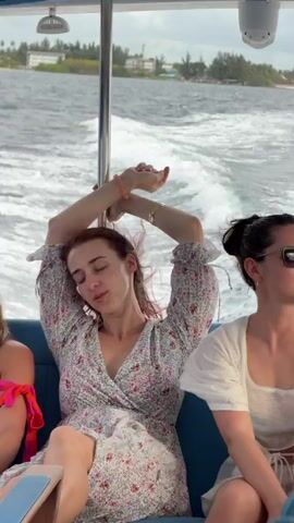 The video shows three women seated on a moving boat. The woman in the center wears a floral print dress and has her arms raised above her head. The woman to her right is wearing sunglasses and a white top, and she is talking to the woman in the middle. The woman to the left is wearing a pink and orange top. The boat is moving on the water, and land is visible in the background.