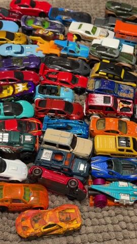 The video begins with a close-up of a collection of toy cars scattered on a brown, textured rug.  The view then widens to show a yellow and light green parakeet walking on the rug, coming into view from the left side of the frame. The bird walks towards the toy cars. The rug the bird and cars are on is partially positioned on a grey, faux marble surface. The bird takes a few steps and looks down at the cars.