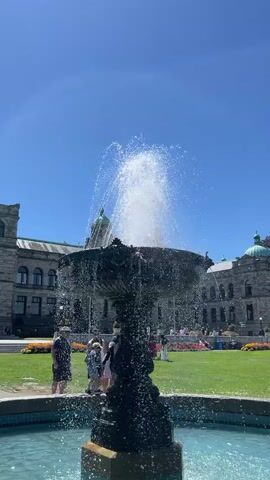 A close-up shot focuses on a decorative water fountain in the foreground, with water spraying upwards and outwards, creating a misty effect. The fountain is ornate, with dark, textured detailing. In the midground, a patch of vibrant green grass is visible, with several people casually strolling or standing. Behind the grass and people, a large, historic-looking building dominates the background. This building features a grey stone facade, arched windows, and prominent green domes. The sky above is a clear, bright blue, indicating a sunny day. The overall scene suggests an outdoor public space, possibly a park or plaza adjacent to a significant government or public building.