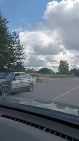 The video shows a view from inside a car driving on a paved road. The sky is blue with white fluffy clouds. Trees and houses are visible on the sides of the road. There are road markings on the asphalt. A traffic sign is visible in the distance. The interior of the car, including the dashboard and windshield wipers, is also visible. The video appears to be taken during the daytime.