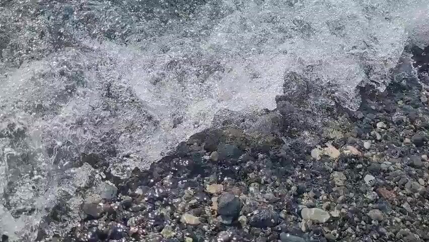 The video captures a close-up, first-person perspective of shallow, clear water flowing over a rocky seabed. The water is slightly agitated, creating ripples and a shimmering effect due to sunlight reflecting off the surface. The seabed is composed of various-sized pebbles and small stones, predominantly dark gray and black, with interspersed lighter-colored rocks. The water appears to be moving from the bottom of the frame towards the top, indicated by the way the light and pebbles shift. There are no people, animals, or man-made objects visible in the frame. The environment is natural, likely a beach or a shallow riverbed.