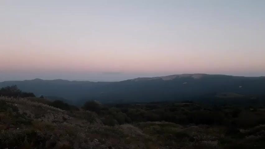 A panoramic video captures a dusky landscape. The sky transitions from a pale pink at the horizon to a pale blue above. In the middle ground, a range of dark, rolling hills stretches across the frame. The foreground features a slope covered in dry grass and scattered bushes, some appearing as silhouettes against the dim light. The overall impression is of a serene, natural environment at twilight.