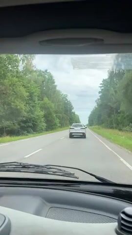 The video shows a view from inside a car looking forward through the windshield at a two-lane asphalt road. Trees line both sides of the road, forming a canopy overhead in some areas. The sky is cloudy. A white car is visible driving in front of the camera car. The dashboard of the camera car is visible in the foreground.