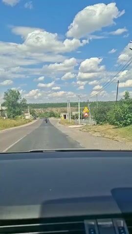 The video begins from the perspective of a driver looking out through the windshield of a car. The road ahead is paved and appears to be a two-lane road with a clear dividing line. The driver is traveling on a sunny day, as indicated by the bright blue sky filled with fluffy white clouds. On the right side of the road, there are utility poles with wires running between them. Further down the road, there are several traffic signs. One sign is triangular with a red border and a symbol of a pedestrian crossing. Another sign shows a circle with a red border and a horizontal line, indicating a speed limit, with the number 30 visible inside the circle. There's also a smaller triangular sign with a red border and a symbol of a pedestrian crossing. On the left side of the road, there are trees and some buildings visible in the distance. A motorcycle is seen traveling in the same direction as the car, further ahead on the road. The landscape beyond the road is hilly and covered with vegetation. The camera is static and does not move.