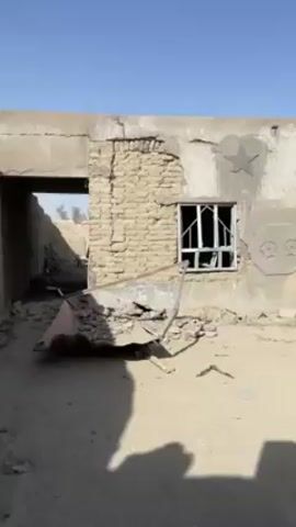 The video shows an exterior shot of a partially destroyed building on a sunny day. A portion of the wall near the left is crumbling, revealing brick underneath. The building also has one window with broken glass, and three concrete reliefs of a star, an angled stripe, and a pair of octagons. Debris litters the ground in front of the building. Several men enter the frame. One man carries a brown bundle, and another man carries a white metal ladder.