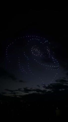 A drone light show at night displays a ghostly figure, possibly a character from the animated series, Rick and Morty. The sky is dark, but silhouetted clouds are visible in the bottom of the frame. Some unseen people stand below the drone light show.