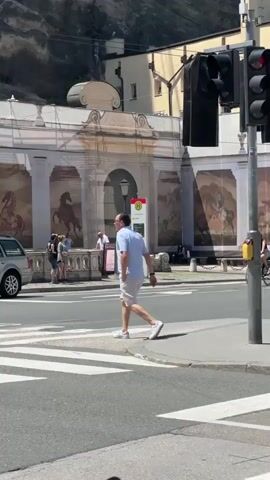The video captures a street scene with people and vehicles. In the foreground, a person on a motorcycle is partially visible. A silver car is on the left, and a zebra crossing is in the center. A man in a light blue shirt and beige shorts walks across the zebra crossing, looking to his left. Behind him, a woman on a bicycle rides to the right. Further back, a man in a red shirt and white shorts walks across the zebra crossing, wearing a red cap and headphones. Another man in a white shirt and sunglasses is in the middle ground, facing right. In the background, there is a building with murals of horses. Traffic lights are visible. A sign with a red diagonal line through silhouettes of two people is prominent. The scene appears to be a public square or street in a European city on a sunny day.
