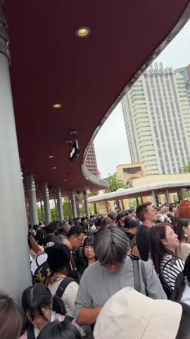 The video shows a very dense crowd of people, mostly young adults, gathered in what appears to be an entrance or waiting area of a theme park. In the foreground, a large, gray cylindrical pillar extends from the ground to a red/maroon ceiling that has recessed lighting.

The crowd fills the entire frame from foreground to background, with individuals standing shoulder-to-shoulder. Many are looking forward, some are looking down, and a few are looking around. Personal items such as hats, backpacks, and various clothing styles are visible.

In the mid-ground and background, the iconic "UNIVERSAL" globe of Universal Studios is partially visible on the left, confirming the location as Universal Studios Japan in Osaka. To the right of the globe, a light-colored building with arches and what looks like a banner with partially visible text, possibly "Breeze Villa," can be seen. Palm trees are also visible behind the crowd, indicating an outdoor or semi-outdoor setting.

The lighting suggests it is daytime, but the sky appears overcast. The people seem to be patiently waiting or slowly moving through a crowded space, characteristic of a queue at a popular attraction. No strong emotions are overtly displayed; the general atmosphere is one of a busy, yet orderly, crowd.