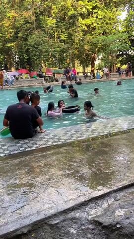 The video shows an outdoor swimming pool with several people swimming and sitting by the edge. In the foreground, a man in a black t-shirt is sitting on the edge of the pool, facing away from the camera, with two children next to him. One child is holding a green and white inflatable ring. Several other people, including adults and children, are in the pool, some with inflatable rings. The pool is surrounded by trees, and there are picnic tables and umbrellas visible in the background. The water in the pool appears clear and blue. The overall atmosphere seems relaxed and recreational.