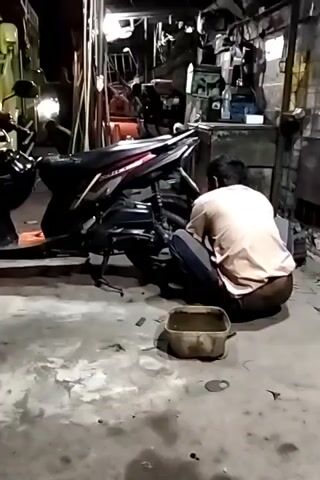 A person is crouched down, working on a motorcycle. The motorcycle is black and has its brake lights on. A yellow truck is parked to the left of the motorcycle. The scene appears to be a workshop or garage. There is a container on the floor in front of the person.