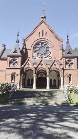 The video opens with a shot of a red brick church with a large rose window and arched entrance. The camera then pans to the right, showing a path leading away from the church, with a woman walking in the distance. The path is lined with trees and bushes, and there is a small fountain in the foreground. The fountain is made of stone and has water flowing from it. The video ends with a close-up of the fountain.