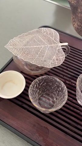 A close-up, overhead shot shows a tea ceremony in progress. A decorative leaf-shaped metal strainer sits atop a clear glass teapot, through which hot water is being poured from an unseen source. Beside the teapot are two small cups, one white and one clear glass with a textured surface. The tea tray is made of dark wood with grooved channels, and a few drops of water are visible on its surface. The overall lighting suggests an indoor setting, possibly with natural light.