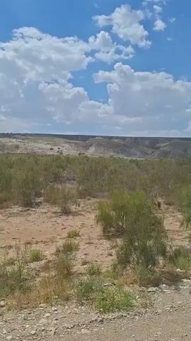 The video shows a barren, dry landscape under a bright blue sky with scattered white clouds. The ground is sandy and rocky, with sparse, dry vegetation. In the distance, there are rolling hills and a dirt road can be seen winding through the terrain. The overall impression is one of an arid, open environment. The camera appears to be moving, possibly from a vehicle, as indicated by the slight shakiness of the footage and the shifting perspective. There are no people or animals visible in the frame.