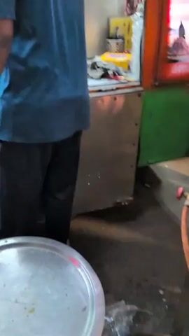 A street vendor in a teal t-shirt stands with his back to the camera, facing a food stall that appears to be set up on a street or sidewalk. Three other male street vendors are visible behind the counter of the food stall. They are preparing and serving food, possibly pizzas or flatbreads, as several round flat food items are seen on metal trays. The stall is brightly lit by fluorescent lights and has a busy atmosphere with people in the background. A large water jug and various other containers are visible on the counter.