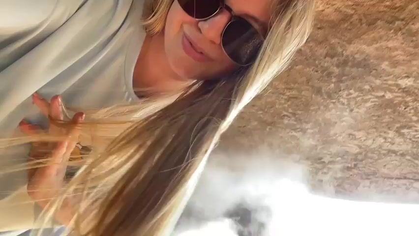 A woman with blonde hair and sunglasses is leaning forward. She is wearing a light blue t-shirt with a cartoon duck logo on the left side. Her hair is long and falls around her shoulders. In the background, there is a waterfall and greenery. The lighting is bright, suggesting it is daytime.