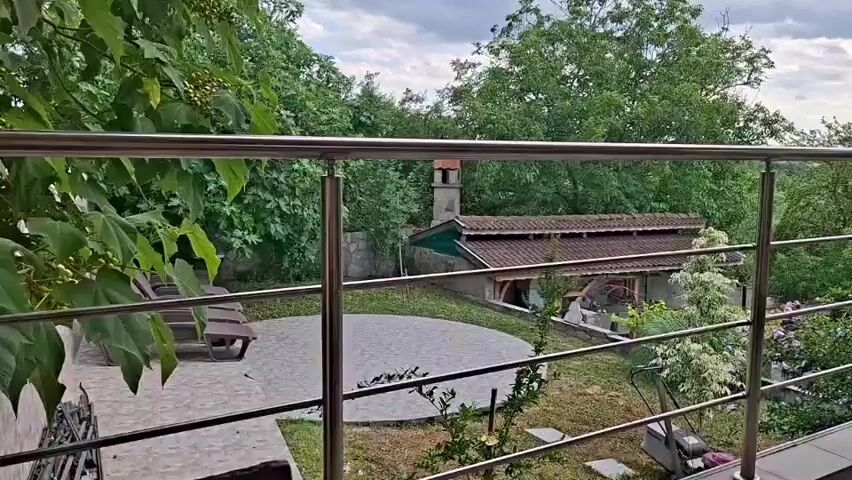 The video pans across a balcony with a metal railing overlooking a backyard. The backyard features a circular patio, lawn chairs, a building with a red tile roof and a chimney, and lush greenery. A chair is visible on the balcony. The sky is cloudy.