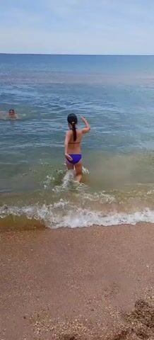 The video shows two people in the shallow ocean near a sandy beach. The person in the foreground is a young female wearing a blue bikini. She is standing with her back to the camera, with her arms outstretched. She is facing a young male who is swimming towards her. The male is shirtless. The water is clear and appears calm, with gentle waves breaking on the shore. The sky is overcast. The overall scene is one of playful interaction in a beach environment.