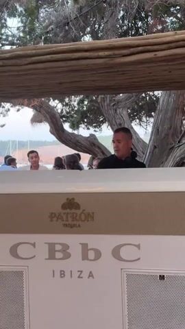 A man in a black shirt stands behind a counter at a venue. The counter has the Patrón Tequila logo and "CBbC IBIZA" written on it. The man is looking down and appears to be interacting with something on the counter. Behind him, other people are visible, and trees and a body of water can be seen in the background. The setting appears to be outdoors, likely a beach club or restaurant.