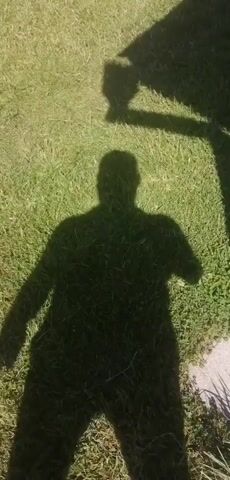 A shadow of a person stands on a grassy surface. The shadow is facing away from the camera. The shadow has one arm raised in the air, with two fingers extended in a peace sign. The shadow's other arm is not visible. The shadow is also partially obscured by another shadow, which appears to be a rectangular object. The sun is shining, casting long shadows. The grass is green and appears to be well-maintained.