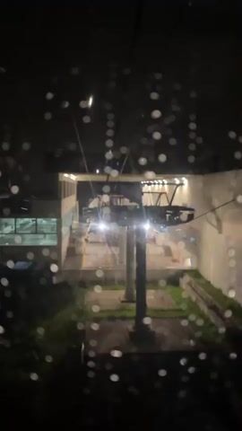 The view from a cable car during rainfall, at night. Through the rain-speckled window, the cable car terminal is seen. The terminal is a modern structure with concrete columns and a well-lit area with a metal frame. The cables supporting the cable car are visible. The overall lighting is dim, creating a contrast between the bright terminal and the dark surroundings. In the background, there's what appears to be vegetation. The camera seems stable, capturing a stationary view of the scene.