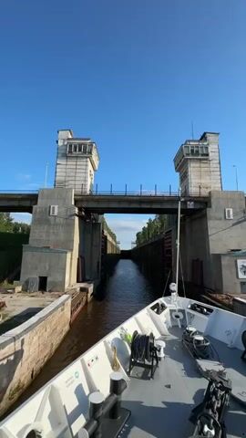 A boat approaches a canal lock. The boat is white with some black trim and appears to be moving forward into the lock. The lock has high concrete walls on either side with two large square towers at the entrance, one on each side. A bridge spans the canal between the towers. The water in the canal is dark brown. The sky is a clear, bright blue with a few wispy clouds. Trees can be seen in the distance beyond the lock. The overall impression is one of approaching a large engineering structure.