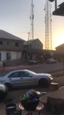 The video shows a street scene in what appears to be a developing country.  In the foreground, a table holds a bowl filled with various metal and plastic containers. Next to it are a few buckets. Behind the table is a parked silver Mercedes-Benz car. Across the street, there's a row of buildings, some with closed storefronts. Several people walk on the sidewalk. A three-wheeled vehicle, a tuk-tuk, drives along the street. In the background, two tall cell towers stand against the sky, where the sun is setting or rising, casting a warm glow.  A few other cars are visible on the road. The scene seems to capture a moment of daily life in this area.