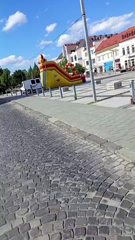 The video pans across a town square on a sunny day. In the foreground, there is a cobblestone street and a paved walkway. Further back, a bright yellow inflatable slide with red and orange accents is set up in the square. Several modern buildings line the square, some with outdoor seating for cafes. A white van is parked in the background. A tall lamppost with multiple lights stands in the middle of the paved area. The sky is blue with scattered white clouds.
