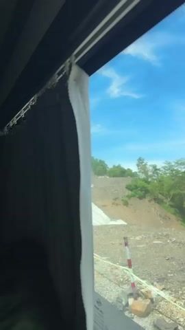 The video shows a view from inside a vehicle looking out a window. A white curtain is partially drawn, covering the left side of the window. Outside, a bright blue sky with scattered white clouds is visible. The landscape appears to be a sloped hillside with patches of green trees and brown dirt. There is a large, white, textured material covering a significant portion of the hillside, possibly a form of erosion control or stabilization. A thin green wire runs horizontally across the lower part of the window. The light suggests it is daytime.