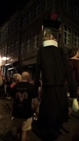 The video shows a street scene at night. In the foreground, a person wearing a black t-shirt with a ram graphic and white shorts is walking away from the camera. To the left, a woman in a pink dress is partially visible. In the center of the frame, two large figures, likely puppets or costumes, are walking down the street. The figure on the left is dressed in a long black robe and a distinctive headpiece. The figure on the right is wearing a maroon tunic. Other people are visible in the background, and the street is lined with buildings that appear to be shops or residences. The ground is paved with cobblestones, and there are streetlights illuminating the scene. The atmosphere appears festive, possibly a parade or festival.