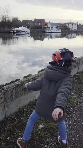 A young boy in a red and black bike helmet, green jacket, jeans, and brown shoes throws rocks into a canal. The canal is lined with a concrete wall and bordered by houses. His bike lays on the ground nearby. The water is calm and reflects the sky and trees.