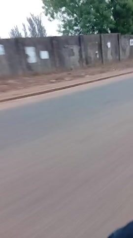 A video captures a street scene from a moving vehicle. The road is asphalt, and alongside it, there is a raised dirt shoulder bordering a concrete wall. On the other side of the wall, trees are visible. As the video progresses, pedestrians in jackets and a helmet are seen walking along the shoulder of the road.  Dilapidated buildings are visible in the background. The overall impression suggests an urban or semi-urban environment.