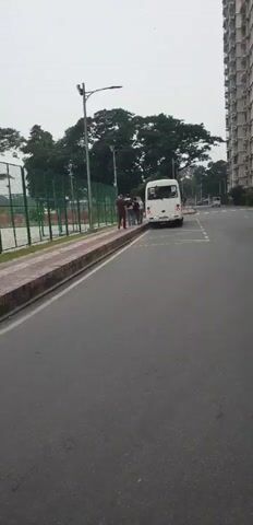 A white bus is parked on the right side of the road. Several people are standing near the bus. A person in a yellow vest is standing further down the road. A tennis court is visible behind a green fence on the left side of the road. A sidewalk with brick detailing runs along the left side of the road. Tall buildings and trees are in the background. The sky is overcast.