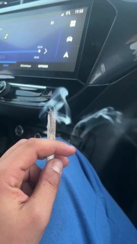 The video, shot from inside a moving car, shows a person holding a lit joint, with smoke wafting around the interior of the vehicle. The dashboard and infotainment screen are visible, displaying navigation information. The camera then pans upwards towards the windshield, showing the highway and the sky outside, which is slightly overcast. The focus is on the joint and the smoke, suggesting the subject is actively engaged in smoking.