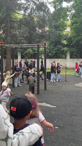 The video captures an outdoor music performance on a playground. In the foreground, two children, a boy wearing a black cap with a red emblem and a girl with long brown hair, are watching the event. Behind them, a group of young people, dressed in casual attire, are performing on stage. Several of them are playing guitars and one is behind a keyboard. A small audience of children and adults are gathered to watch. The setting appears to be a park or schoolyard, with trees and a fence in the background. The ground is a dark, rubberized surface, typical of playgrounds. The performance is likely taking place during the daytime, with overcast skies suggesting a cloudy or possibly humid day. The overall atmosphere seems to be relaxed and joyful, as indicated by the engaged audience and the energetic performance. No visible text is present in the video. The location is identified as Dmitrov, Russia.