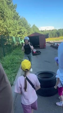 A woman in a black t-shirt and grey pants is putting on a white and red racing helmet. She has a blue hairnet on her head. She is standing on a paved area with two young girls in front of her. One girl wears a yellow hat and a pink t-shirt with purple shorts, and the other wears a purple hat and a white t-shirt with pink pants. In the background, there is a red go-kart with the number 8 on it inside a small wooden structure. To the right, there are stacks of black tires. The environment appears to be an outdoor go-kart track, with trees and green vegetation surrounding the area. The sky is blue.