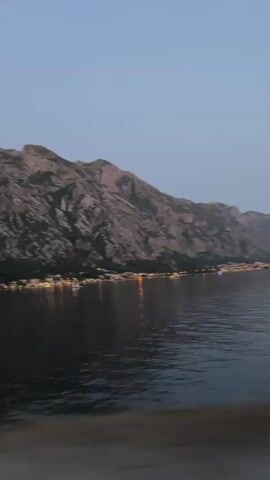 The video displays a scenic view of a body of water with mountains in the background. The water is dark and reflects the distant lights of a town or village situated along the coastline at the base of the mountains. The sky is a pale, dusky blue, suggesting either dawn or dusk. The camera appears to be in motion, possibly from a moving vehicle, as evidenced by the slight blur of trees in the foreground during certain frames. There is a small white boat visible in the water. The mountains are rocky and rugged, with patches of greenery. The overall atmosphere is serene and tranquil.