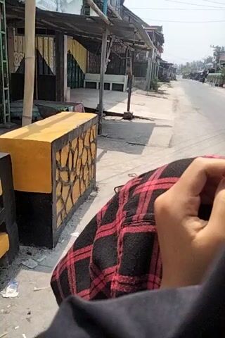 The video shows an outdoor scene, possibly a street or alleyway, with several structures on the left. The structures appear to be shops or small buildings with wooden framing and corrugated metal roofs. Some of the buildings have closed shutters and signs. In the foreground, there is a concrete block with a textured, stone-like pattern painted on it in yellow and black. A person wearing a red and black checkered shirt is partially visible in the upper right corner of the frame. The ground is paved, with some cracks and debris visible. Further down the path, a person is riding a motorcycle. The overall impression is that of a lived-in, possibly semi-urban or rural environment.