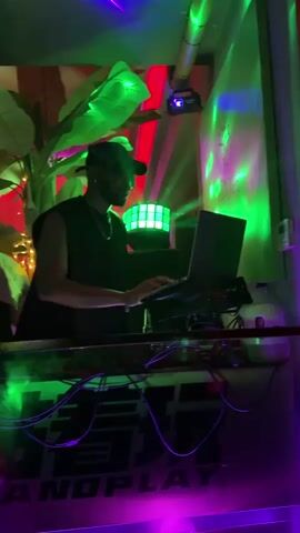 Here is a description of the video:
The video shows a DJ in a club playing music. The DJ is wearing a black sleeveless shirt and a baseball cap and has headphones around his neck. He is standing behind a DJ console and using a laptop. The club is decorated with tropical plants and colorful lights, including red, green, blue, and purple. There are several people in the club, including a few who are dancing. A neon sign that says "Oh My House" is visible in the background. The atmosphere appears energetic and lively.